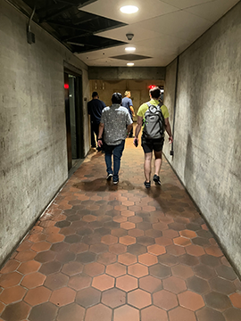 A picture of a pathway in the metro station from the elevator. There are five men in the background of the picture, two of which are walking further away. There are concrete walls on both sides of the picture.